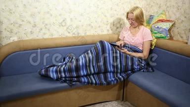 金发女人坐在沙发上用毯子盖着，在平板电脑上玩